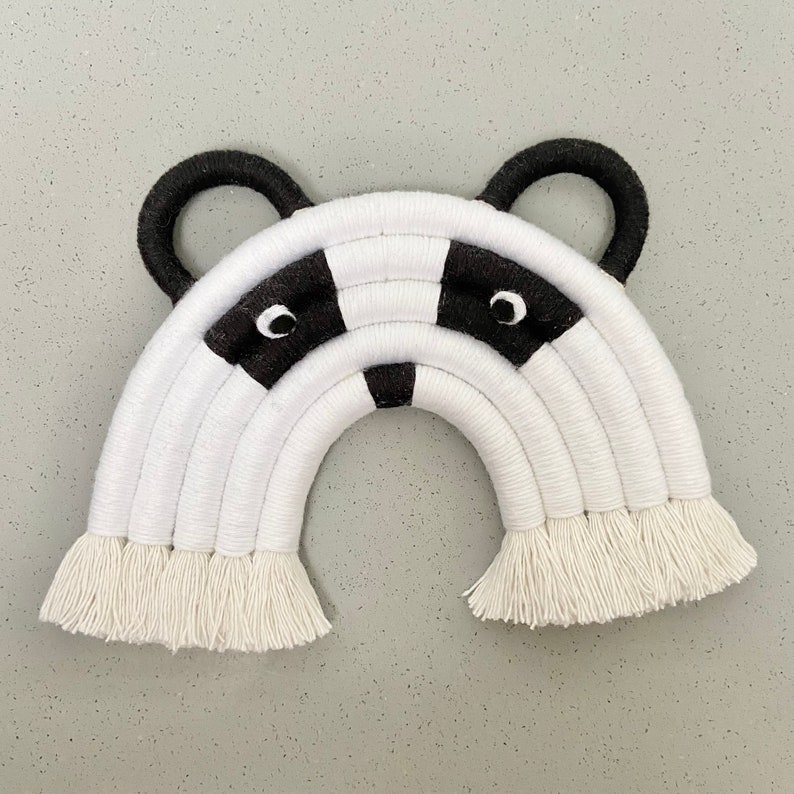 Puede incluir: Colgante de pared de arco&iacute;ris tejido en blanco y negro con una cara de mapache blanco y negro en el centro. El arco&iacute;ris tiene una franja en la parte inferior.