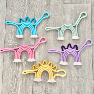 May include: Five colourful rainbow-shaped macrame wall hangings in the form of dinosaurs. The colours are light blue, light green, pink, purple, and yellow.