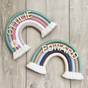 Puede incluir: Dos arcoíris de hilo de colores con letras de madera que deletrean los nombres "OTILIE" y "EDWARD". Los arcoíris están decorados con flecos blancos y una margarita blanca.