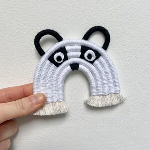 Puede incluir: Colgante de pared de arco&iacute;ris tejido en blanco y negro con cara de mapache. El mapache tiene orejas negras, nariz negra y dos ojos grandes blancos.