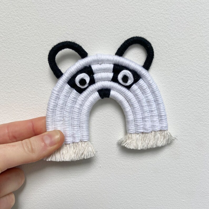 Puede incluir: Colgante de pared de arco&iacute;ris tejido en blanco y negro con cara de mapache. El mapache tiene orejas negras, nariz negra y dos ojos grandes blancos.