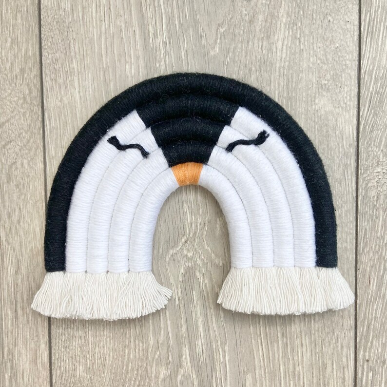 Op de afbeelding: Een zwart-witte regenboog wanddecoratie met een franje rand. De regenboog heeft een zwart-wit pingu&iuml;n gezicht in het midden.