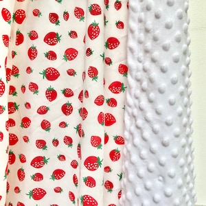 May include: White fabric with a repeating pattern of red strawberries on a white background. The fabric is next to a white textured fabric with raised dots.