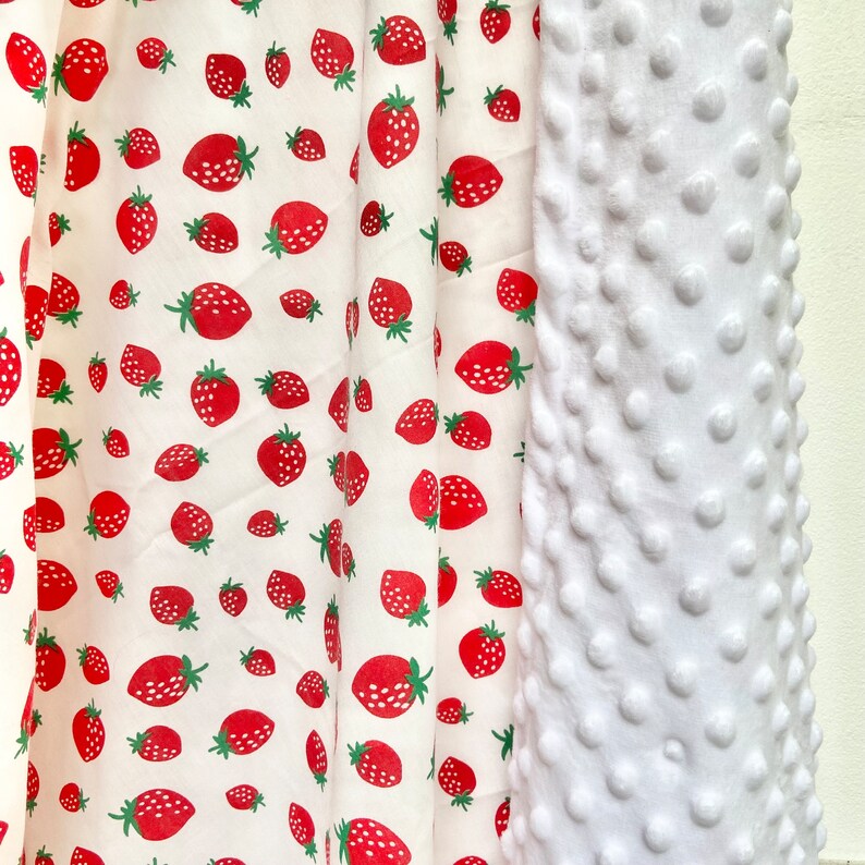 May include: White fabric with a repeating pattern of red strawberries on a white background. The fabric is next to a white textured fabric with raised dots.