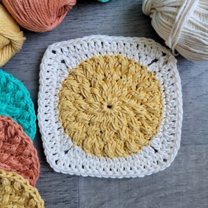 May include: A crocheted square with a yellow center and a white border. The square is surrounded by balls of yarn in various colors, including orange, green, and white.