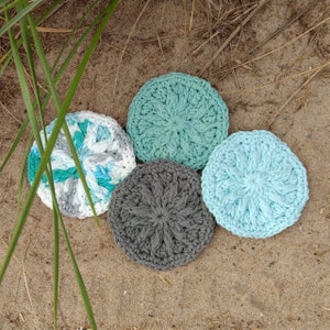 May include: Four crocheted coasters in different colours and patterns. Two are light blue, one is grey, and one is white with blue and green accents.