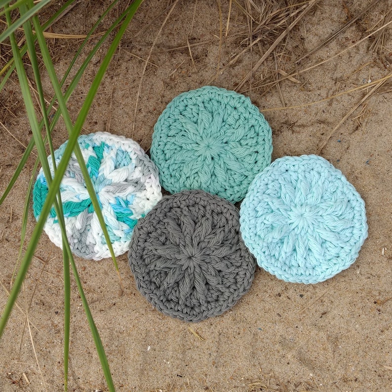 May include: Four crocheted coasters in different colours and patterns. Two are light blue, one is grey, and one is white with blue and green accents.