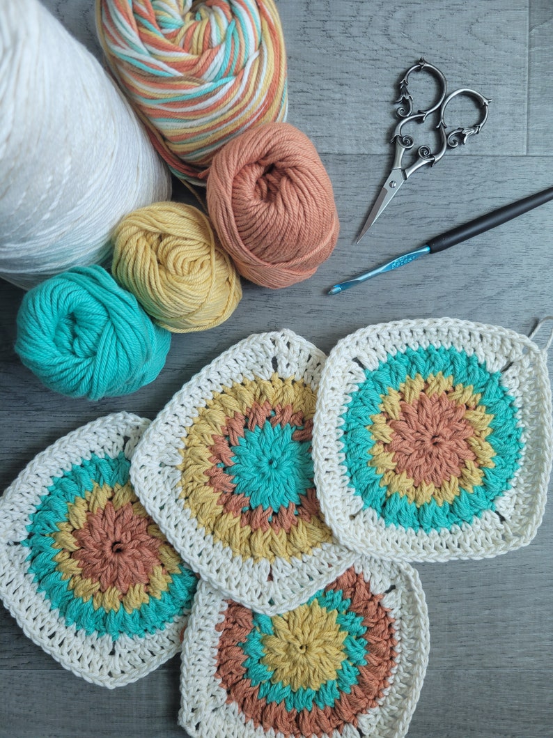 May include: Four crocheted squares in shades of white, teal, yellow, and orange. The squares are arranged in a staggered pattern, with the largest square in the center. There are also balls of yarn in various colors, a crochet hook, and a pair of scissors.