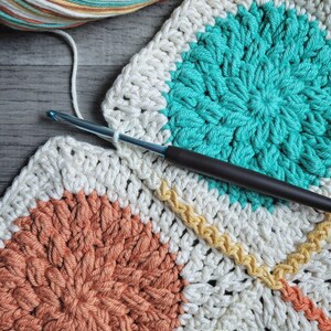 May include: A close-up of a crocheted blanket with a variety of colors, including white, teal, orange, and yellow. The blanket is being worked on with a crochet hook.