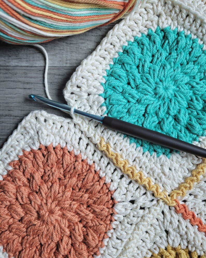 May include: A close-up of a crocheted blanket with a variety of colors, including white, teal, orange, and yellow. The blanket is being worked on with a crochet hook.