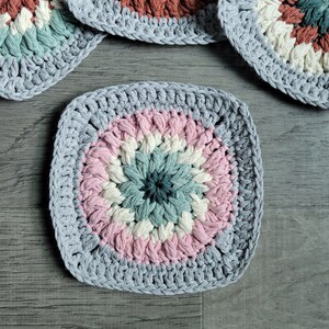 May include: Four crocheted square coasters with a multi-colored center design. The coasters are made with a light gray yarn and feature a pink, white, and green center.