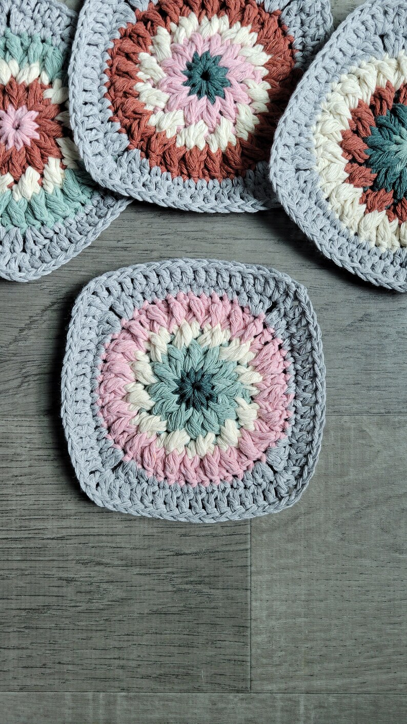 May include: Four crocheted square coasters with a multi-colored center design. The coasters are made with a light gray yarn and feature a pink, white, and green center.