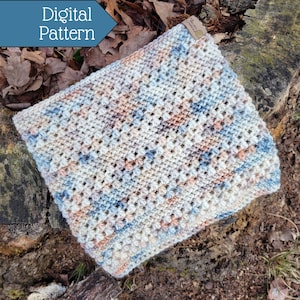 May include: A crocheted cowl in a light blue, beige and peach colour scheme. The cowl is made with a textured stitch and has a subtle geometric pattern. The cowl is lying on a tree stump with leaves and dirt in the background.