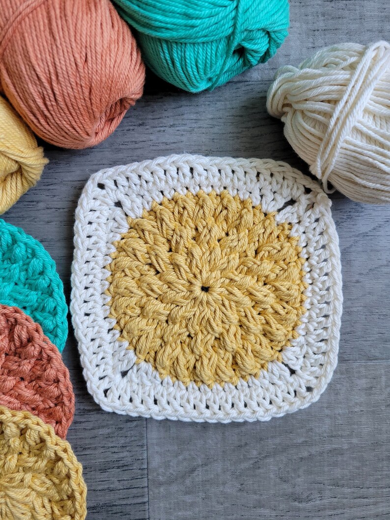 May include: A crocheted square with a yellow center and a white border. The square is surrounded by balls of yarn in various colors, including orange, green, and white.