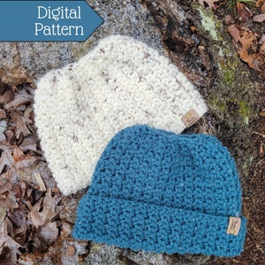 May include: Two crocheted hats, one white and one teal blue, are lying on a bed of brown leaves and moss. The hats have a textured stitch pattern and are both adorned with a small brown leather tag.