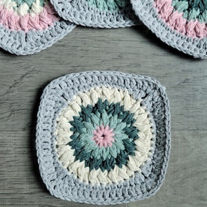 May include: Four crocheted squares with a colorful floral design in shades of pink, white, green, and brown. The squares are made with a grey yarn and have a textured, raised surface.