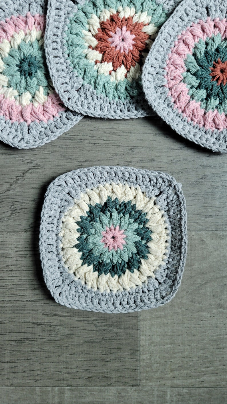 May include: Four crocheted squares with a colorful floral design in shades of pink, white, green, and brown. The squares are made with a grey yarn and have a textured, raised surface.