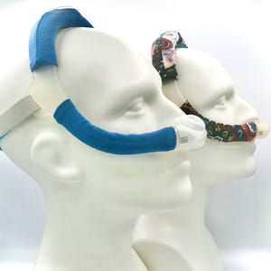 May include: Two white mannequin heads display CPAP mask accessories. One has a blue fabric cover, the other a patterned fabric cover. The accessories are designed to fit around the mask's headgear and tubing.