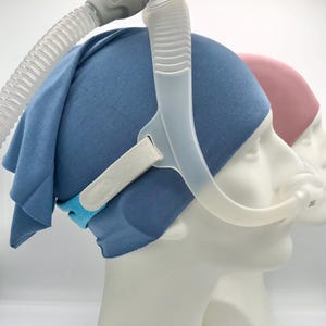 Peut inclure: Un bandeau en tissu bleu porté sur un masque nasal CPAP avec un tube en plastique transparent et un connecteur gris. Le masque comporte une sangle élastique blanche et une bande en tissu bleu. Un second mannequin porte un bandeau rose en arrière-plan.