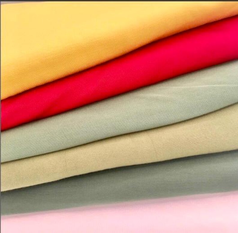 May include: A stack of colorful fabric samples in shades of yellow, red, green, and pink. The fabric appears to be a smooth, woven material, possibly cotton or linen, with a slight sheen. The layers are neatly arranged, showcasing the different colors.