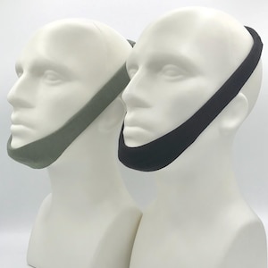 May include: Two chin straps worn on white mannequin heads. One strap is solid olive green and the other is solid black. These fabric bands wrap around the chin and extend upward to the top of the head, providing a secure fit for facial support.
