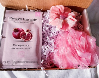 May include: A pink gift box filled with lavender-colored shredded paper. The box contains a pink facial scrub, a pink hair scrunchie, and a facial mask packet with a pomegranate image and the text "Revive the skin. Day by Day Facial mask Pomegranate 라떼 리바이브 더 스킨 마스크".