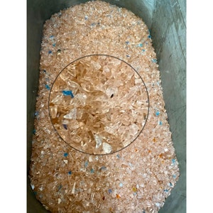 May include: A close-up of a large container filled with translucent, light brown plastic flakes. The flakes are mostly clear with a slight amber hue. Some of the flakes have a blue tint.