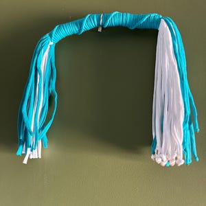 Puede incluir: Un tapiz de macramé de color turquesa y blanco con cuentas blancas en la parte inferior.