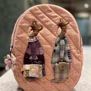 Könnte beinhalten: Ein rosa gesteppter Rucksack mit zwei gestickten Bären, die kleine Taschen halten. Die Bären tragen Kleidung und haben braunes Fell. Der Rucksack hat einen Reißverschluss und einen kleinen Blumenanhänger.