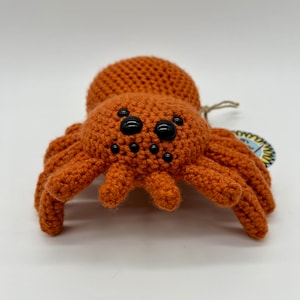 May include: A crocheted orange spider toy with black eyes. The spider has eight legs and is sitting on a white surface.