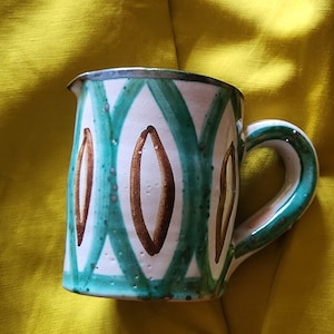 Può includere: Una tazza in ceramica bianca con un motivo geometrico verde e oro. La tazza ha un manico ed è appoggiata su una superficie gialla.