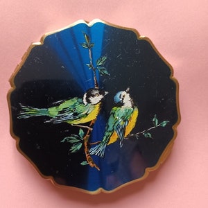 Peut inclure: Un compact rond, bleu et noir avec une bordure dorée présente deux oiseaux jaunes et verts perchés sur une branche. Les oiseaux se font face et ont des yeux noirs.