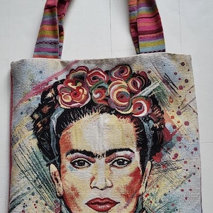 Könnte beinhalten: Tragetasche mit einem Frida Kahlo-Porträt. Die Tasche hat einen gewebten Griff mit einem bunten Muster. Das Hauptbild ist eine detaillierte Darstellung von Kahlo's Gesicht, geschmückt mit einem Blumenkranz. Der Hintergrund zeigt abstrakte Pinselstriche.