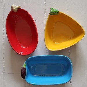 Pode incluir: Três pratos de servir de cerâmica com a forma de vegetais. Os pratos são vermelho, amarelo e azul. O prato vermelho tem a forma de um pimentão, o prato amarelo tem a forma de um pimentão e o prato azul tem a forma de uma berinjela.