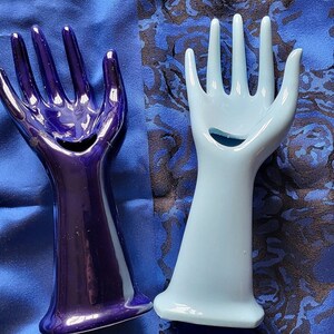 Può includere: Due porta anelli in ceramica a forma di mano. Uno è di colore blu scuro e l'altro è di colore blu chiaro.