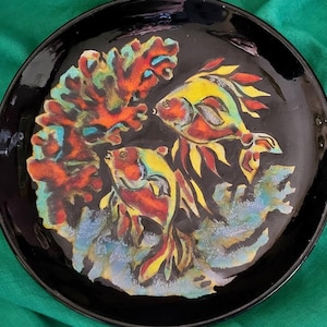 May include: A black ceramic plate with a hand-painted design of coral and fish. The coral is red, orange, and yellow, and the fish are yellow, red, and blue.