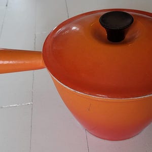 Può includere: Una pentola in ghisa smaltata arancione con coperchio e manico. La pentola ha una forma arrotondata e un pomello nero sul coperchio. Il manico è sul lato sinistro.