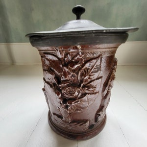 May include: A brown ceramic jar with a lid. The jar features a floral design and is decorated with a raised pattern. The lid is made of metal and has a black knob.