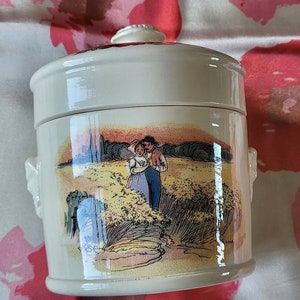 Könnte beinhalten: Eine weiße Keramikdose mit Deckel und zwei Henkeln. Die Dose ist mit einer gemalten Szene eines Paares in einem Weizenfeld verziert.