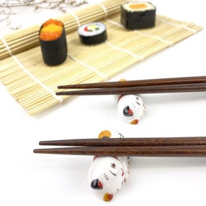 May include: Two pairs of brown chopsticks resting on white ceramic cat-shaped chopstick rests. The cats are laying down with their heads resting on the chopsticks.