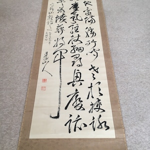 May include: A traditional Japanese scroll painting with black ink calligraphy on a beige background. The scroll features a long vertical line of characters in a flowing script. The characters are written in a traditional style and are likely to be a poem or a quote.