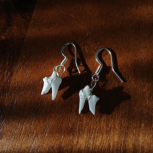May include: A pair of unique earrings featuring white, tooth-shaped pendants. Each tooth is wrapped with silver wire and suspended from a silver fish hook ear wire. The earrings are displayed on a wooden surface.