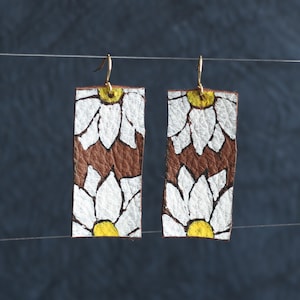 May include: A pair of brown leather earrings with a white daisy pattern and yellow centers.