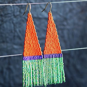 Puede incluir: Un par de pendientes de cuentas con un diseño triangular. La parte superior es naranja, pasando a una banda azul, y luego a flecos verdes y blancos. Los pendientes cuelgan de ganchos dorados.