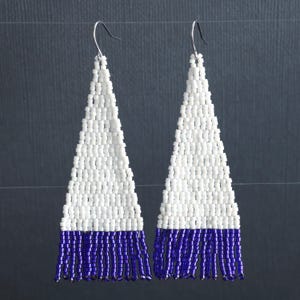 Pode incluir: Um par de brincos longos, brancos e roxos. Os brincos são triangulares e têm uma franja de contas roxas na parte inferior.
