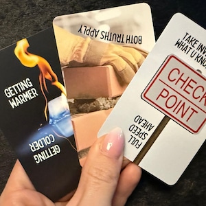 May include: Three tarot cards are held in a hand. The cards have text and graphics. One card says "Getting Warmer" and "Getting Colder" with a flame and ice graphic. Another says "Both Truths Apply" with a bricklaying graphic. The third says "Check Point".