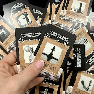May include: A selection of black and tan tarot cards, each displaying chess-related imagery and text. The cards feature illustrations of chess pieces and phrases such as "Queen Me" and "Death". The cards are held in a hand.
