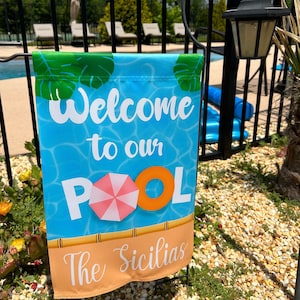 Peut inclure: Un drapeau de piscine rectangulaire avec un fond d'eau bleu et les mots "Welcome to our Pool" en blanc. Il présente un parapluie rose et blanc et une bouée de sauvetage orange. Le bas a une bannière beige avec le nom de famille.