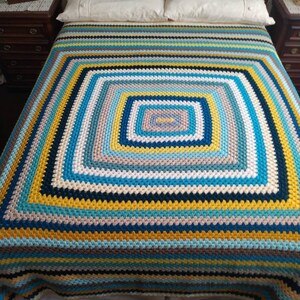Puede incluir: Una gran manta de ganchillo multicolor con un patrón cuadrado en el centro. La manta está hecha con rayas de hilo azul, amarillo, blanco y gris.