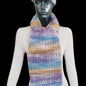 Puede incluir: Una bufanda de ganchillo multicolor con un gorro de pompón. La bufanda está hecha con una variedad de tonos de azul, morado, marrón y beige. El gorro es de color morado claro con un pompón marrón.